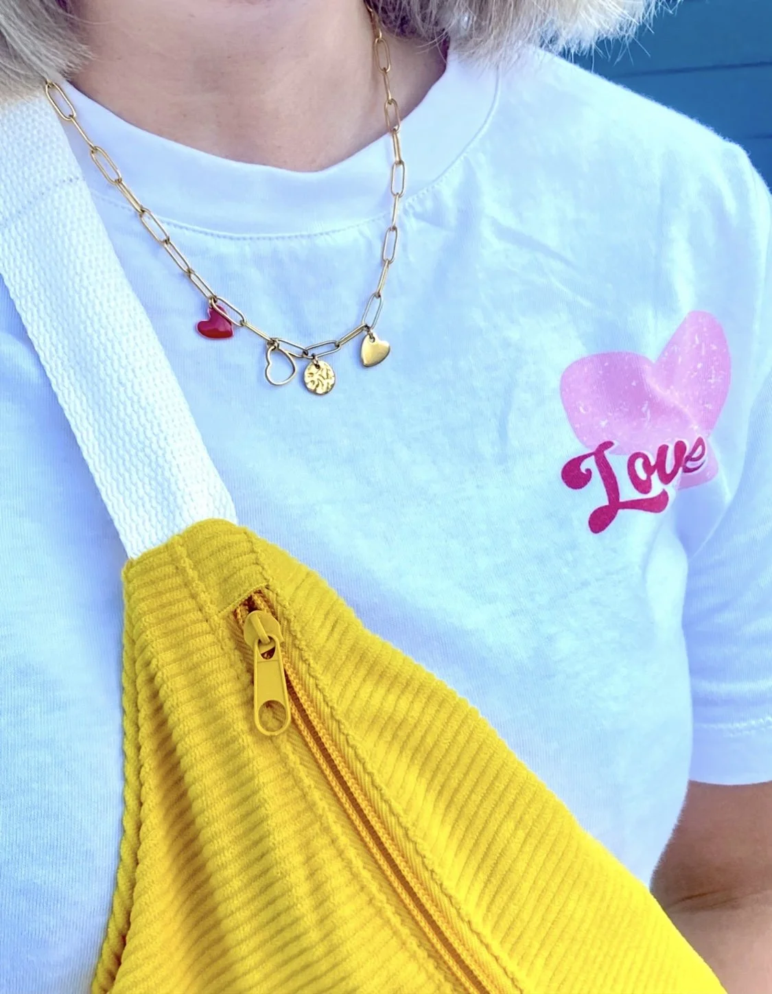 Une personne porte un t-shirt blanc avec un dessin de cœur rose et le mot « Love » écrit en rouge. Elle porte également un collier avec plusieurs pendentifs, dont un cœur rouge, un cercle avec un motif, un cœur doré et un autre pendentif. Elle a un sac banane jaune en velours côtelé avec une fermeture éclair jaune. La personne porte aussi une chemise ou une veste avec une épaule marron clair ou crème.