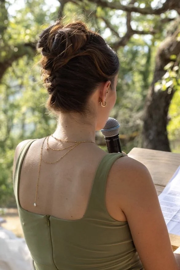 Une femme vue de dos, parlant dans un microphone, avec un chignon coiffé élégamment, portant un haut vert et des bijoux dorés, en extérieur avec des arbres en arrière-plan.