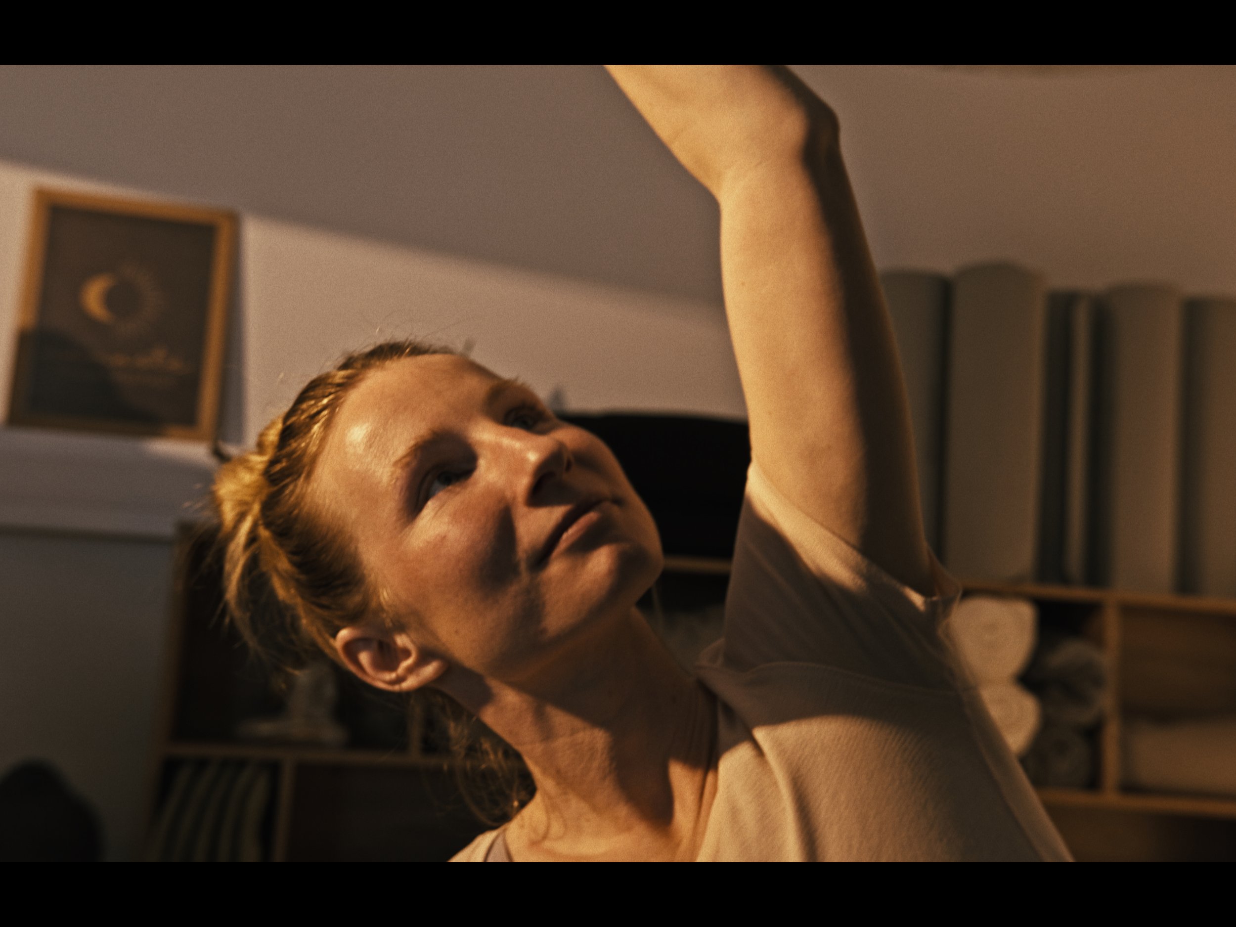 A woman with red hair and light skin is stretching her arm up in a room with bookshelves and framed pictures on the wall in the background.