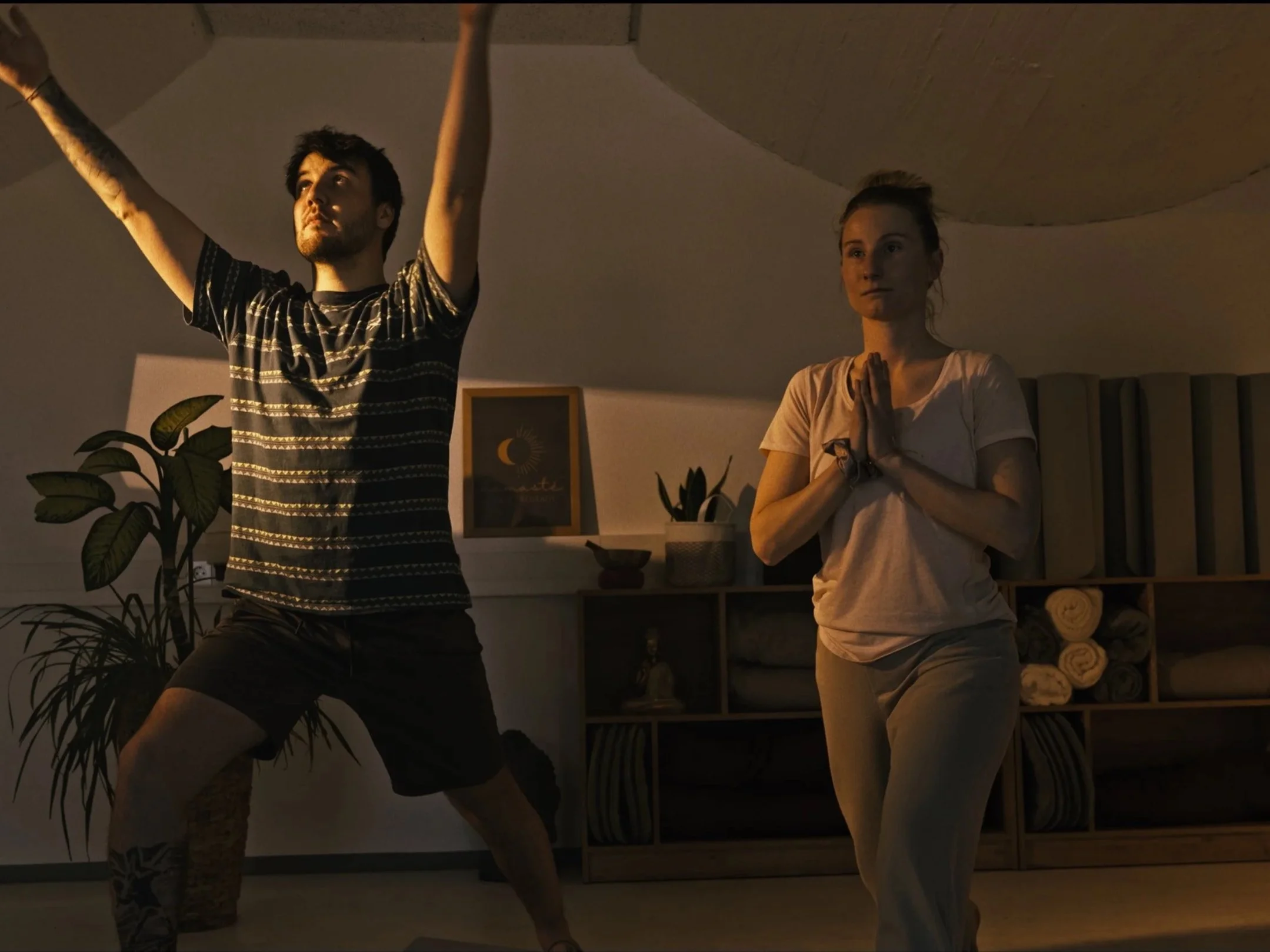 A man and woman practicing yoga in a dimly lit indoor space, with the man stretching his arms upward and the woman standing in a prayer position with hands together in front of her chest.