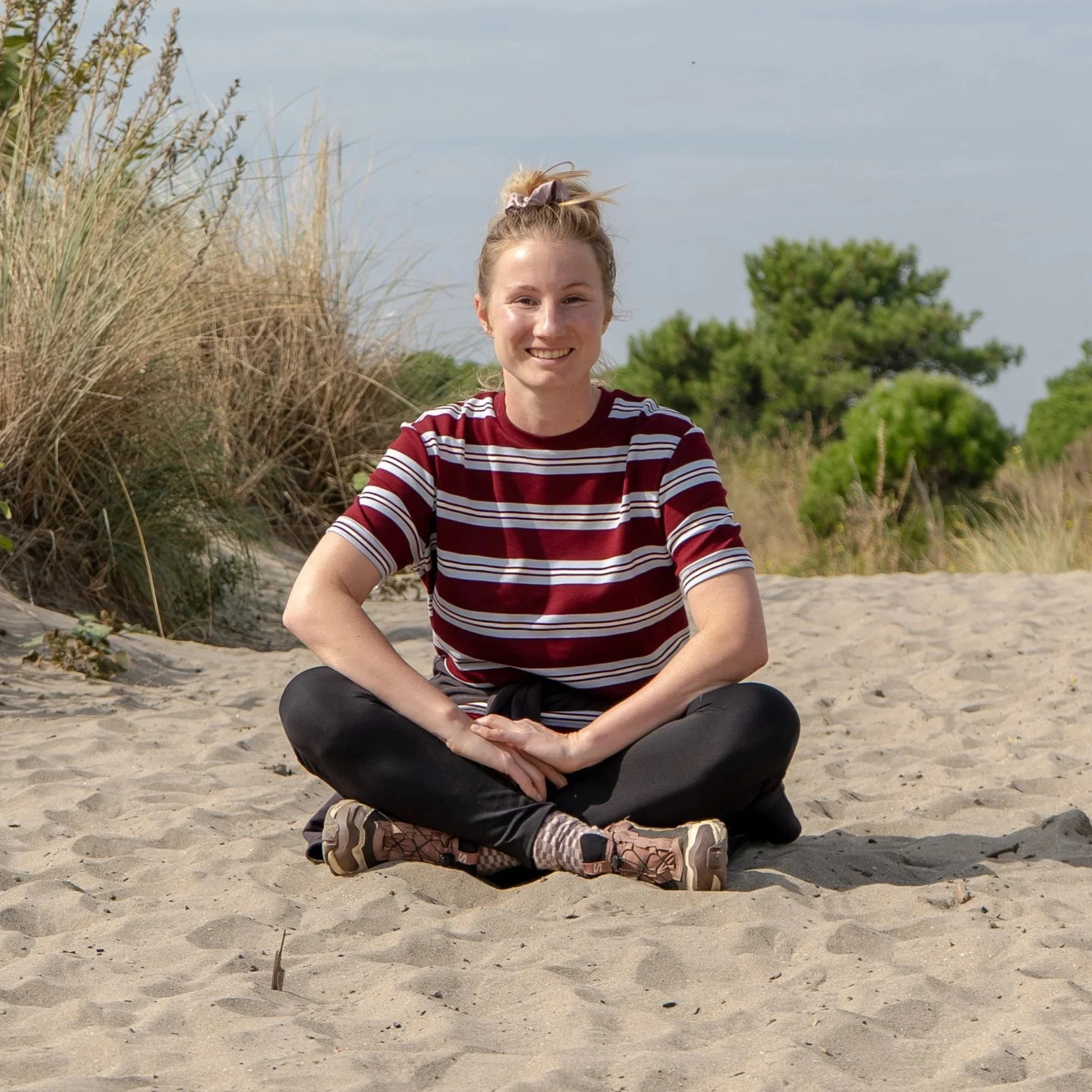 A smiling young woman sitting cross-legged on a sandy beach, wearing a red and white striped t-shirt, black pants, and hiking shoes, with green bushes and trees in the background under a cloudy sky.