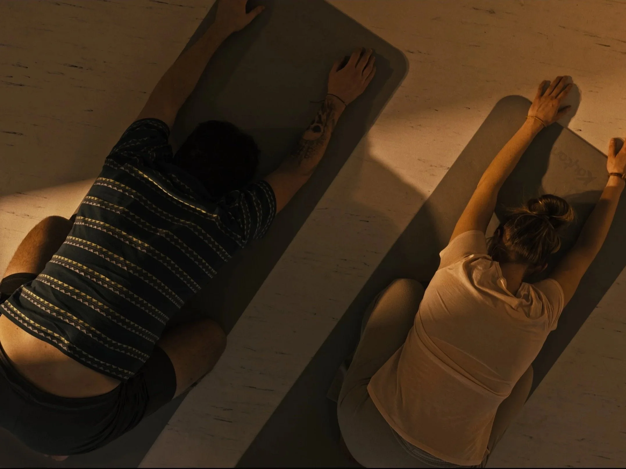 Two people are lying face down on yoga mats, stretching their arms overhead, in a dimly lit room.