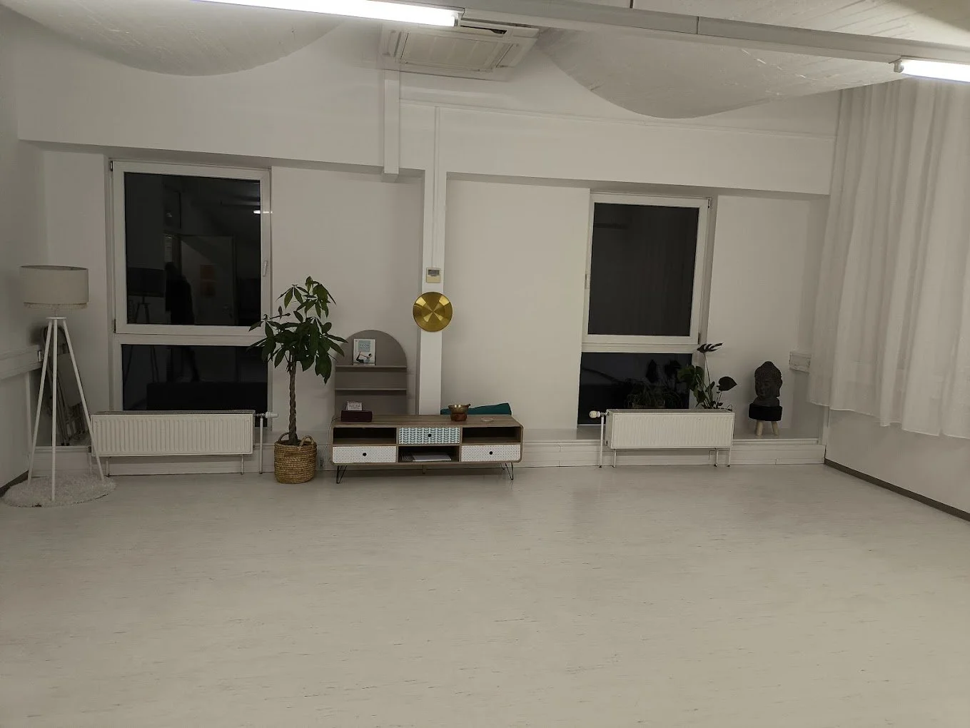 Minimalist living room with white walls, two large windows, and white curtains. Contains a potted plant, a small media console, decorative items, and a black bust sculpture. Ceiling has fabric panels and fluorescent lighting.