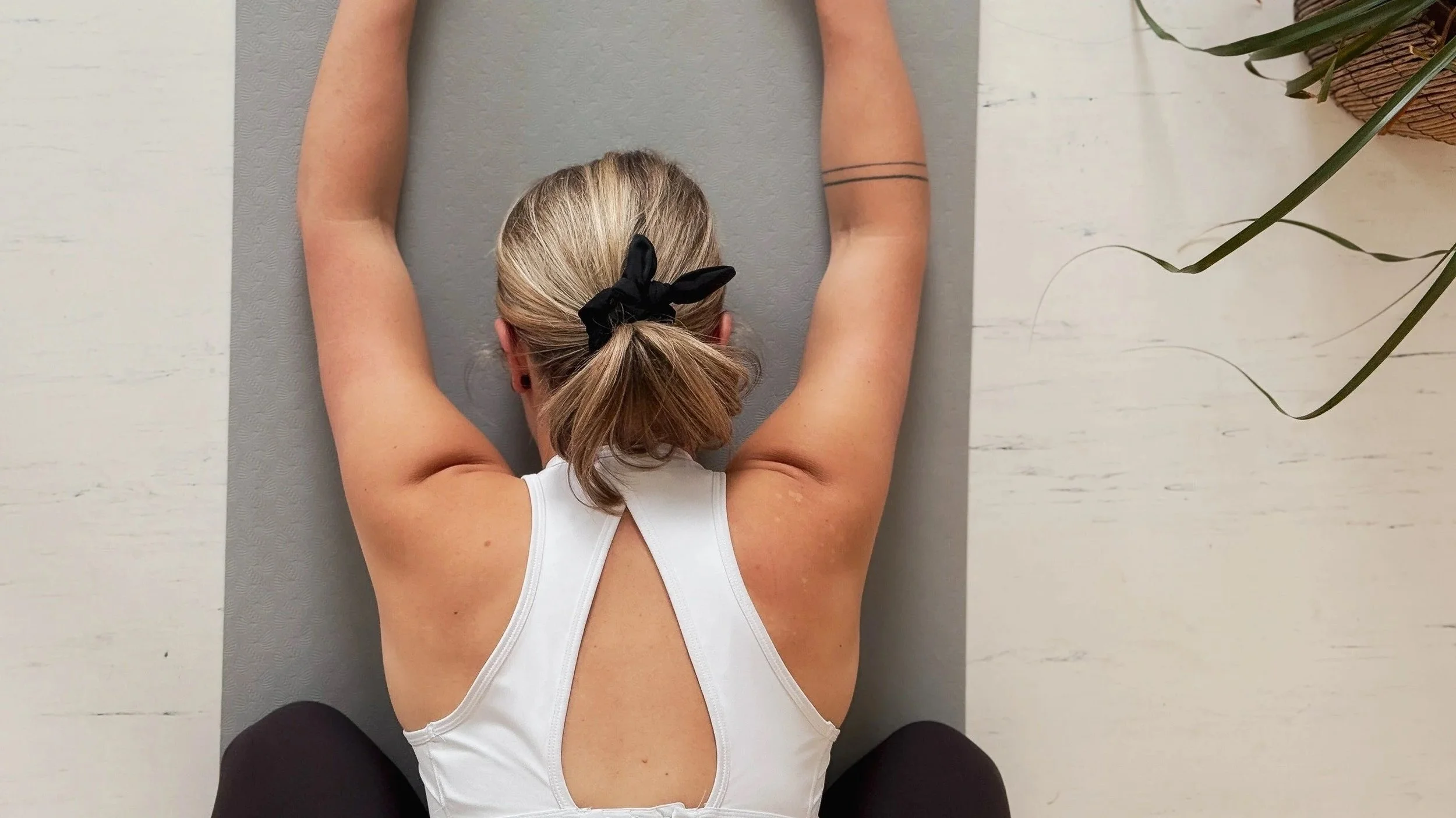 A woman with blonde hair tied with a black bow is practicing yoga on a gray mat, sitting on a white wooden floor near a plant.