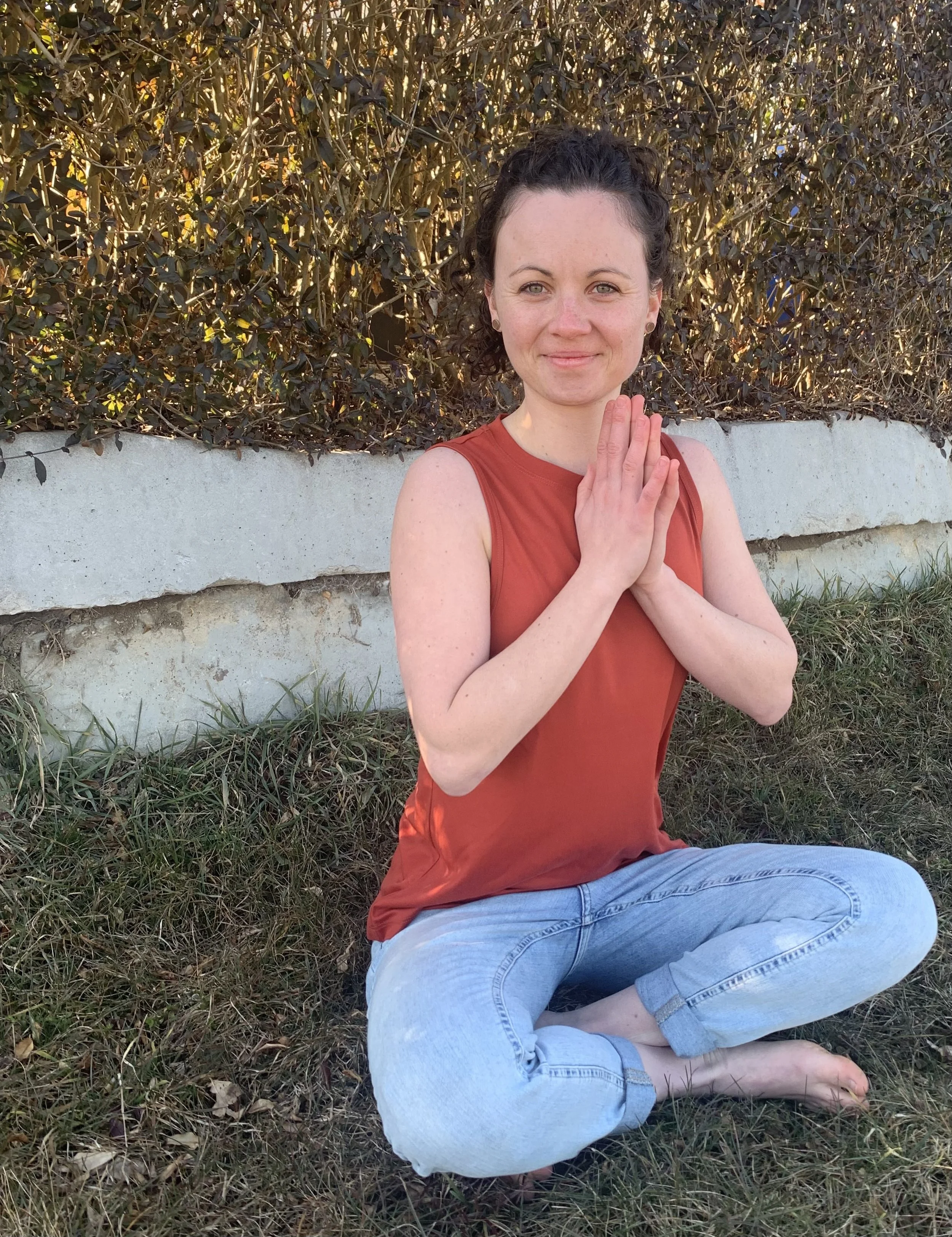 A woman practicing yoga outdoors, sitting cross-legged on grass with hands pressed together in a prayer position, smiling gently at the camera against a background of shrubs and a concrete edge.