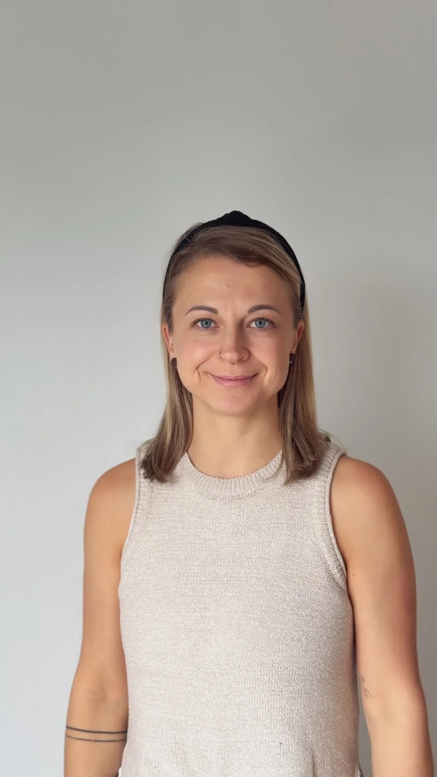 A young woman with blonde hair, blue eyes, and a septum piercing, smiling at the camera, wearing a beige sleeveless top and a black headband, standing against a plain white wall.