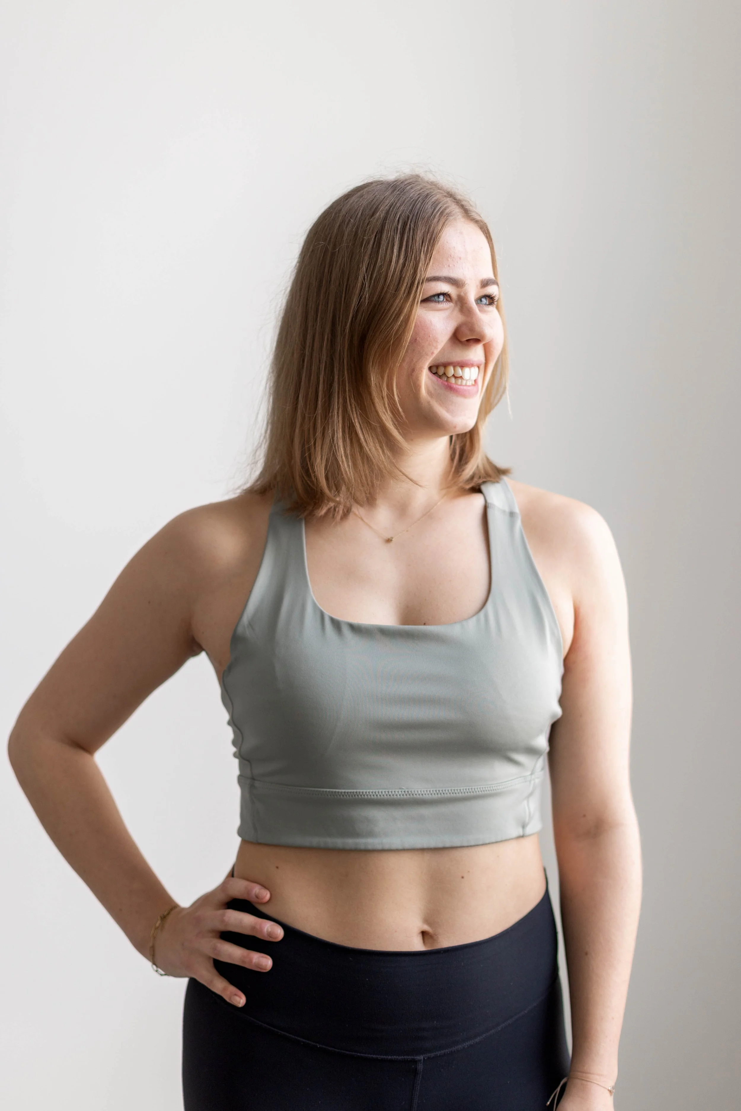 A young woman with shoulder-length red hair and fair skin, smiling and looking to the side, wearing a light gray sleeveless athletic top and black high-waisted leggings, standing indoors against a plain light-colored wall.