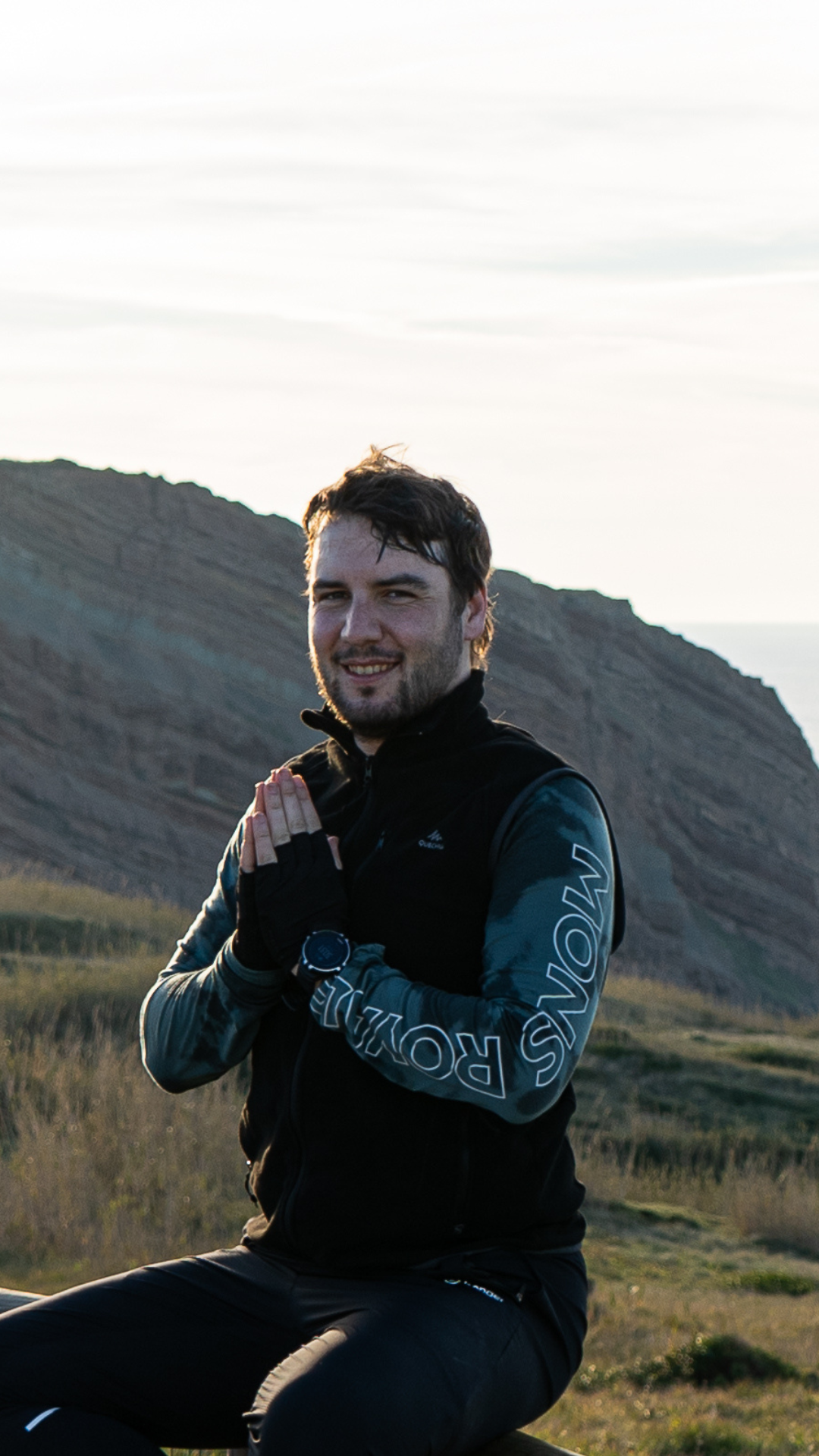 A man sitting outdoors in a mountainous area, smiling and wearing athletic clothing, with hands pressed together in a prayer position near his chest.