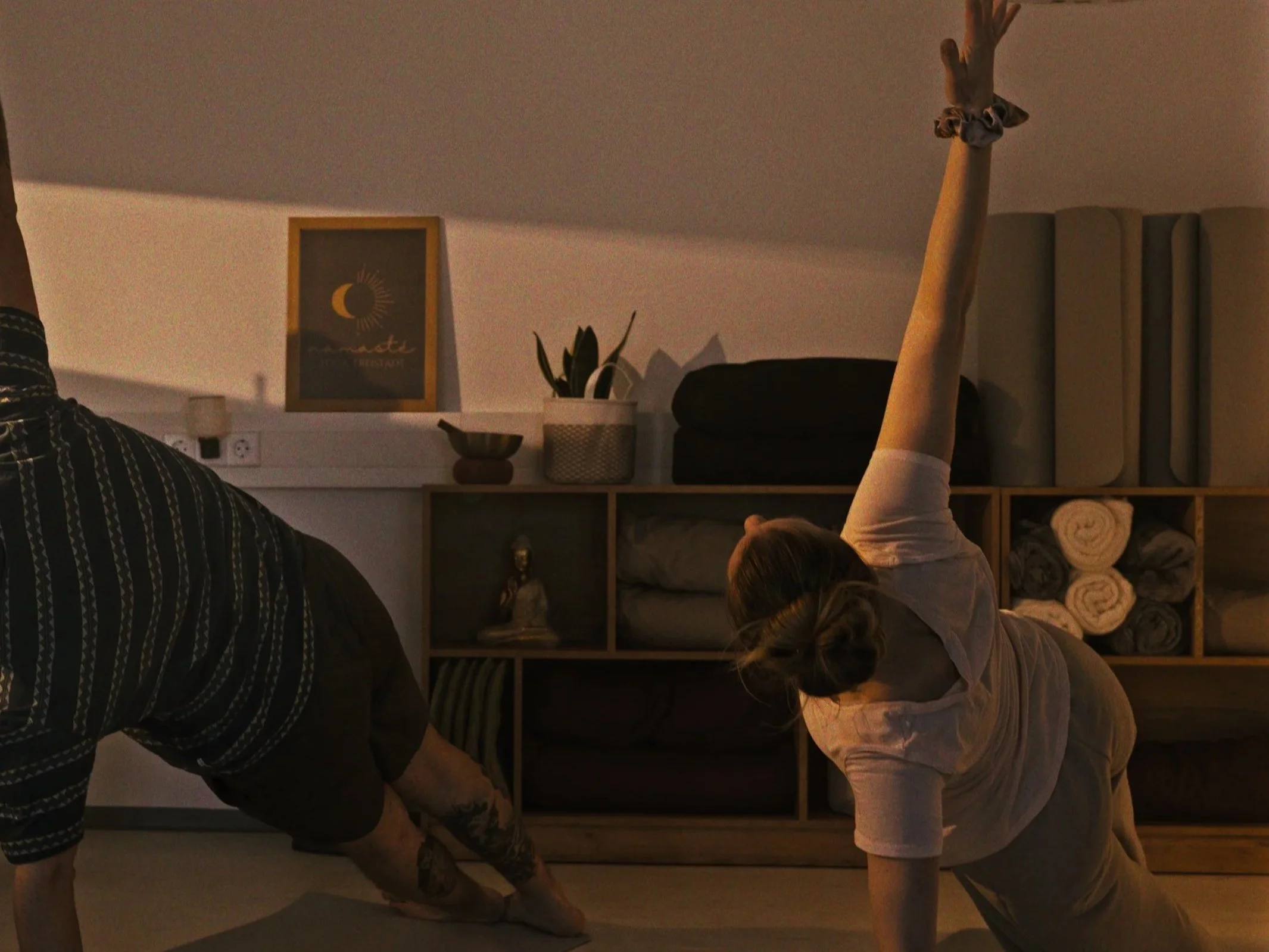 Two women practicing yoga in a cozy room, one reaching upward and the other in a forward bend, with shelves of rolled towels and decorative items in the background.