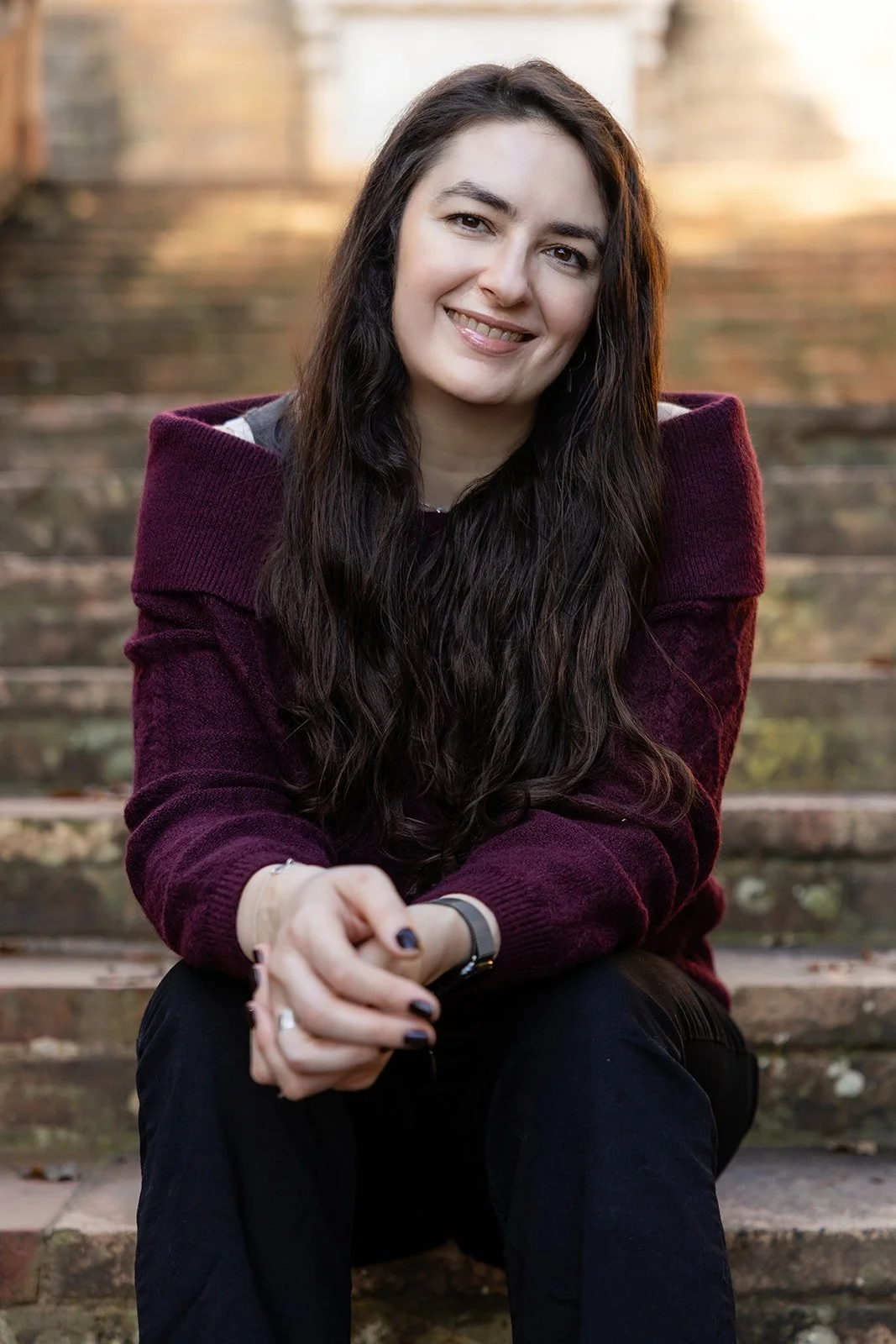 A young woman with long dark hair sitting on outdoor stairs, smiling at the camera, wearing a burgundy sweater and black pants.