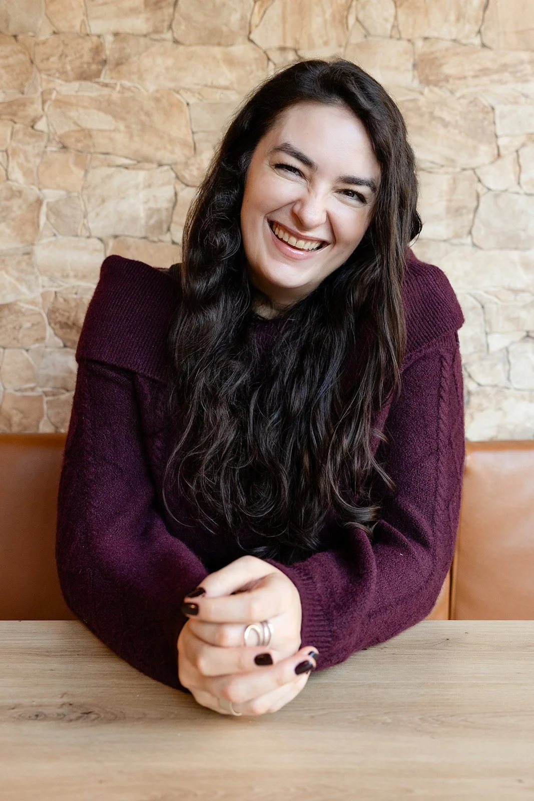 A woman with long dark hair smiling and sitting at a wooden table in front of a stone wall, wearing a dark red sweater.