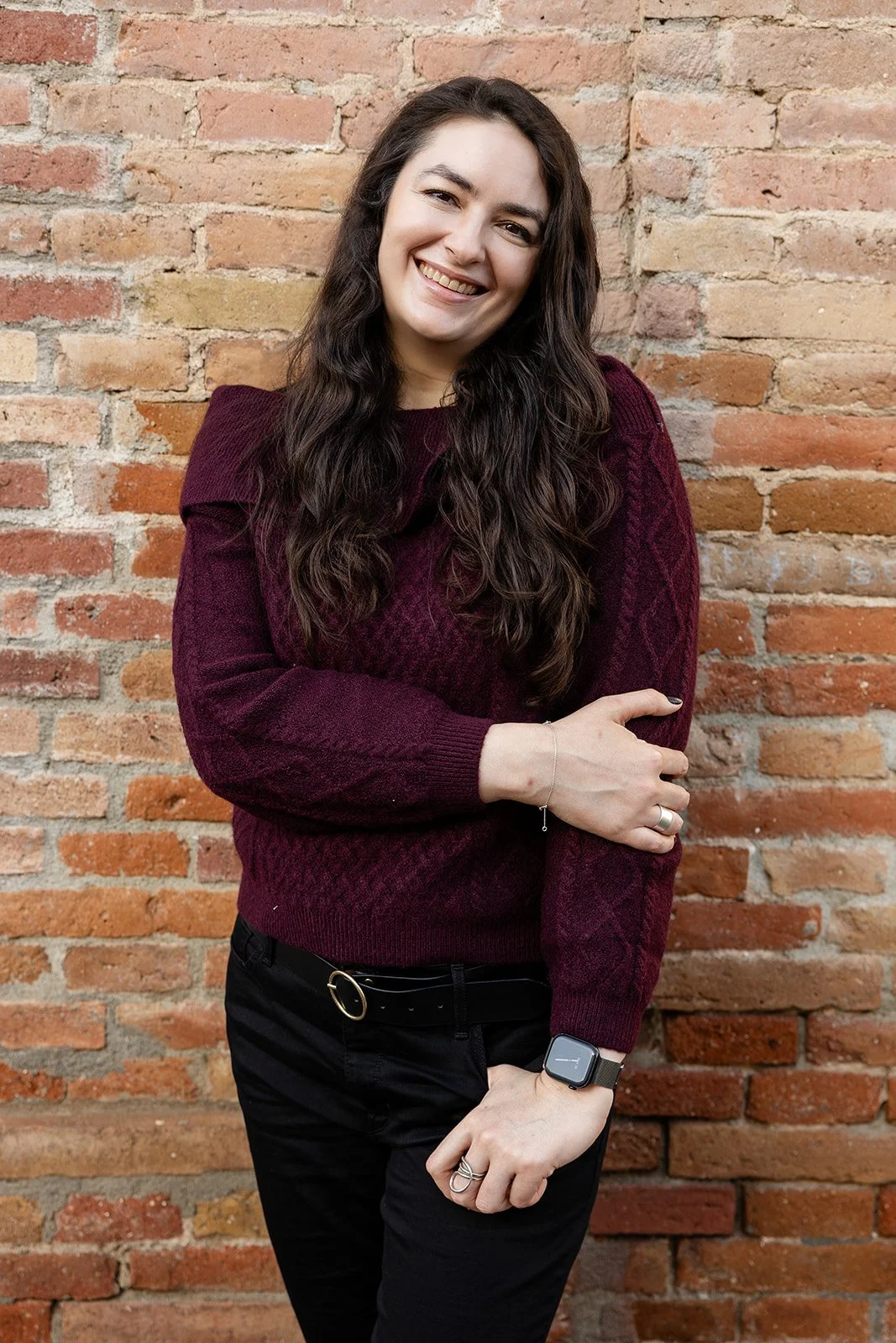 A woman with long, dark hair wearing a maroon sweater and black pants standing in front of a brick wall, smiling and crossing her arms.