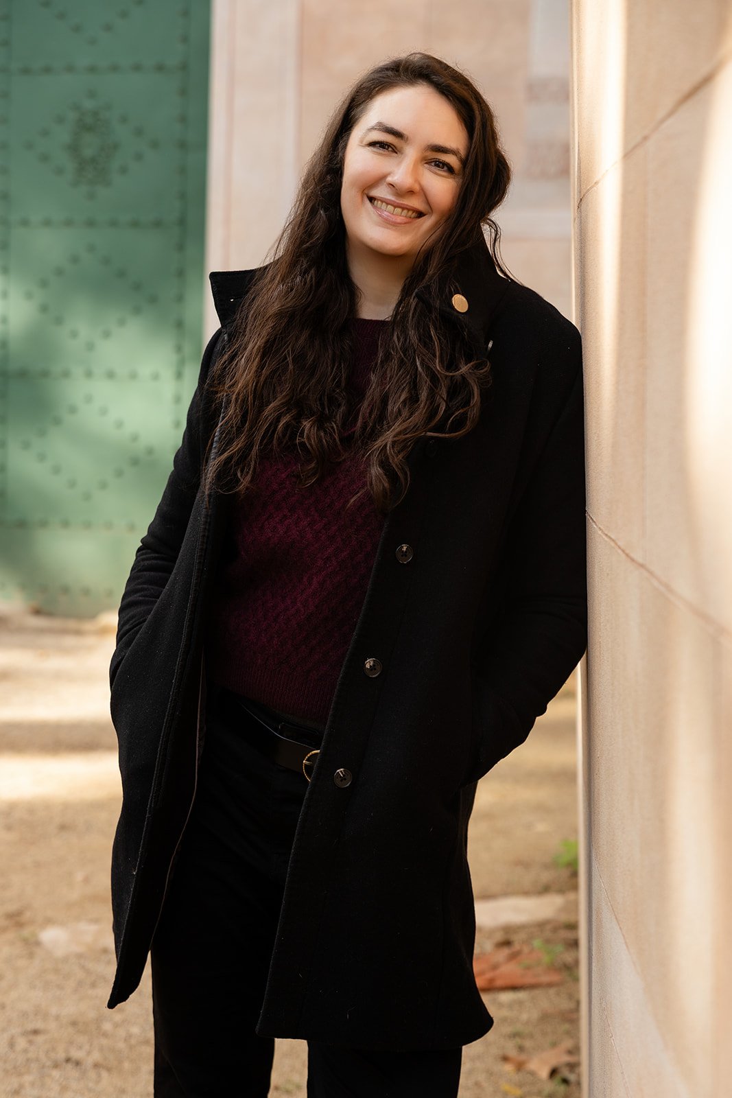 A smiling woman with long, dark, wavy hair leaning against a beige stone wall outdoors.