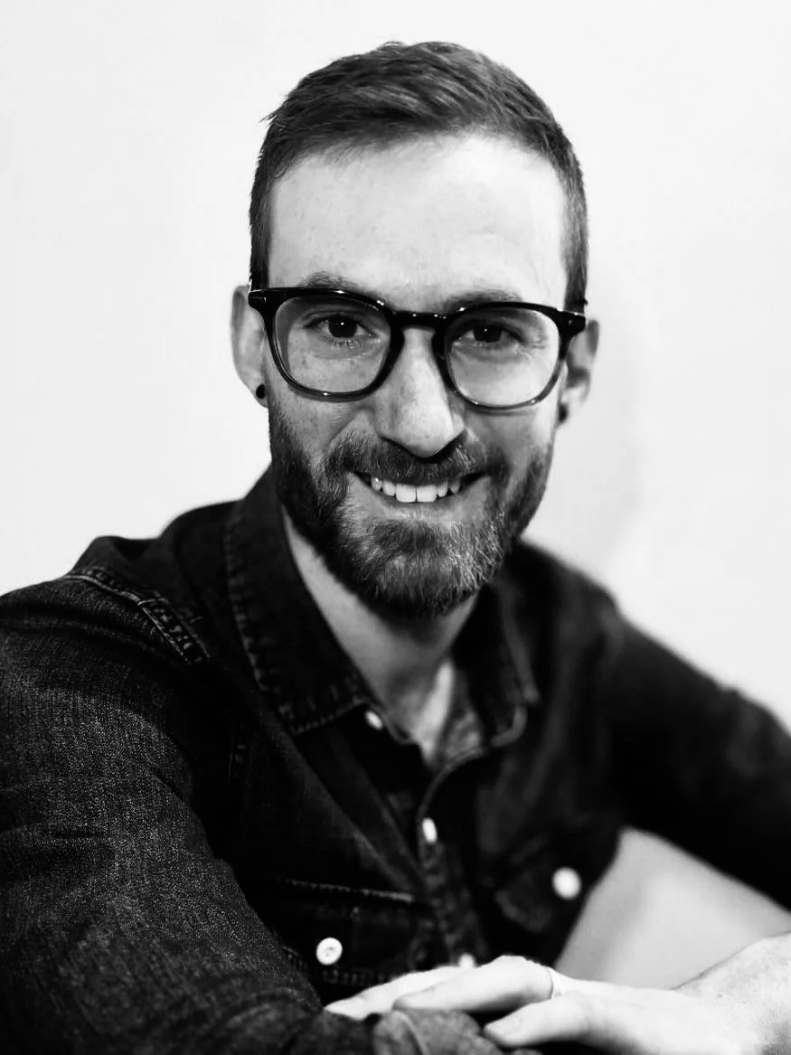 Black and white photo of a young man with glasses, short hair, beard, and a friendly smile, wearing a denim jacket, with arms crossed.