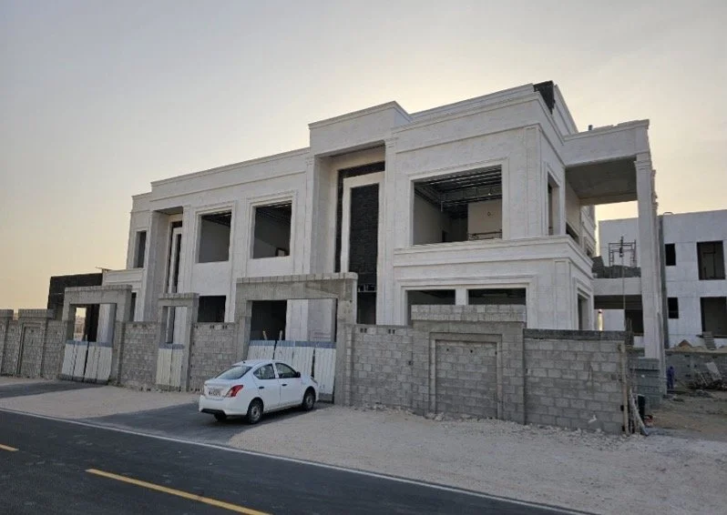 Under-construction modern multi-story house with white exterior walls, large windows, and a car parked in front, surrounded by a gray brick wall.