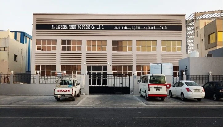Front view of a building with a sign that reads 'AL JAZERA PRINTING PRESS Co. L.L.C.' in English and Arabic, with cars parked in front and a black gate.