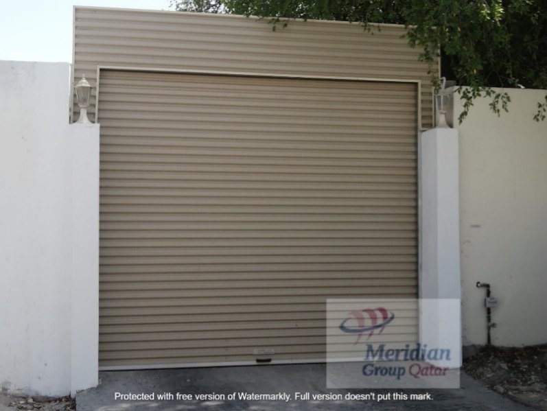 A middle-aged Asian man with dark hair sitting on a beige cushioned bench against a plain beige wall, looking to the side with a contemplative expression, wearing a blue short-sleeved sweater and beige pants.
