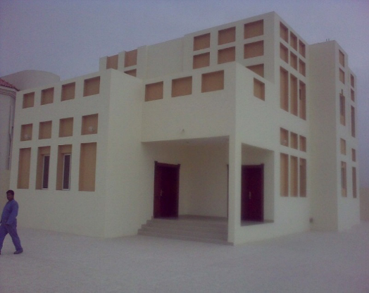 A modern white building with multiple square and rectangular windows, featuring a central entrance with stairs and a person walking nearby.