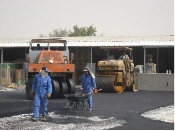 Workers in blue overalls laying fresh asphalt on a road using construction equipment at a construction site.
