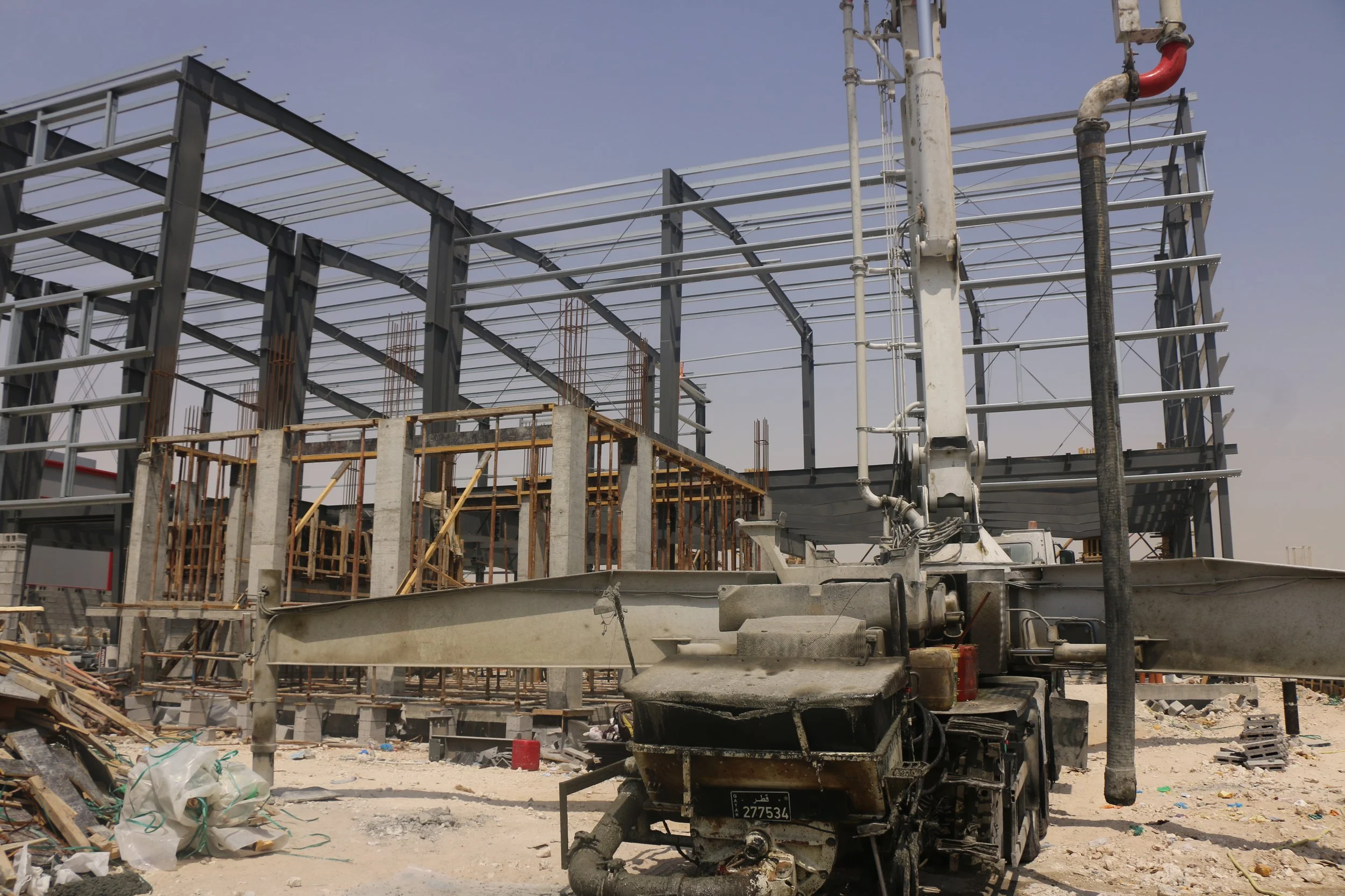 Construction site with a steel framework of a building under construction, a concrete pump truck in the foreground, and construction materials scattered on the ground.