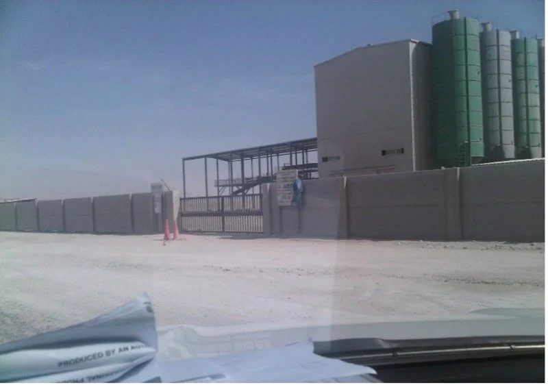 View of an industrial building with green cylindrical tanks, surrounded by a high fence, and a gated entrance from inside a vehicle on an unpaved road.