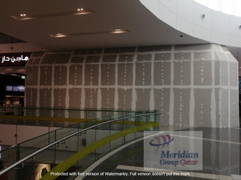 Woman with dark curly hair sitting on a bench in a room with sunlight casting shadows, a cream-colored wall, and a wooden wall panel.
