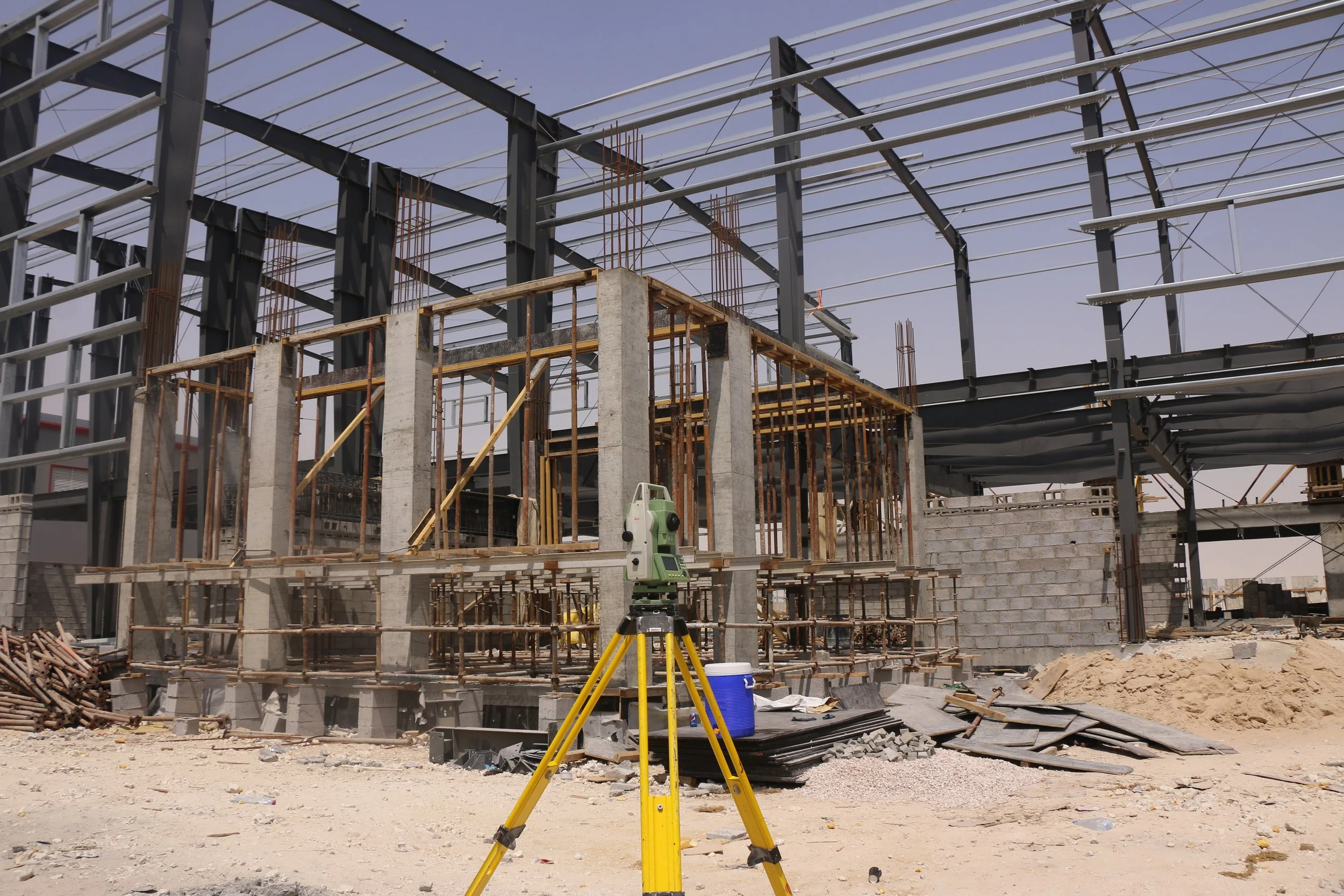 Construction site with steel framing, concrete columns, and wooden scaffolding, including a surveying instrument on a tripod and construction materials scattered on the ground.