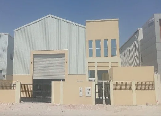 Industrial building with a beige facade, large roll-up garage door, and enclosed yard with a gate and fence under a clear blue sky.