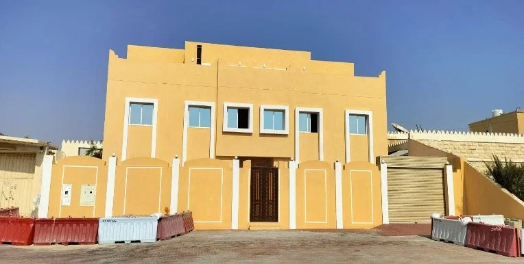Yellow building with multiple small windows and a black entrance door, surrounded by a low wall, with a clear blue sky in the background.