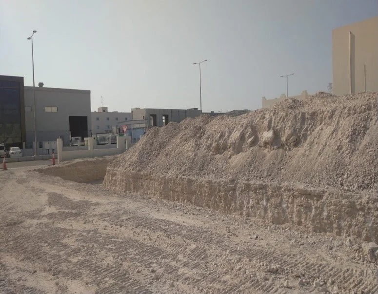 A construction site with a large dirt mound and residential buildings in the background.