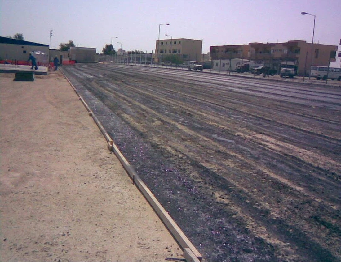 A nearly finished dirt and asphalt racetrack with a concrete curb on the left side, in an urban area with low-rise buildings and parked cars in the background.