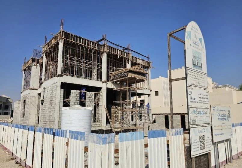 Under construction building with scaffolding and workers, surrounded by a white and blue fence.