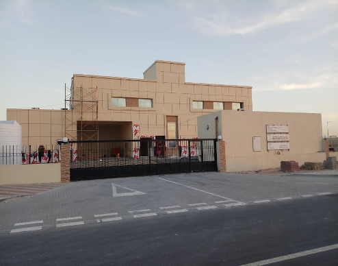 A modern, beige, multi-story building with scaffolding on one side, surrounded by a black gate and a paved sidewalk in front.