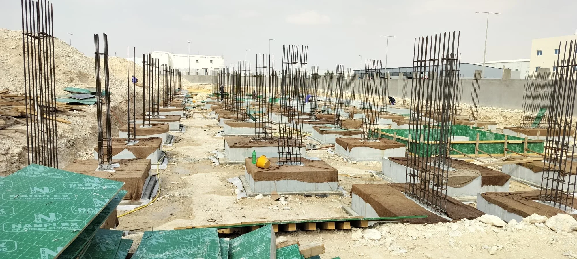 Construction site with foundation pillars and steel rebar for a building, dirt ground, construction materials, and workers in the background.