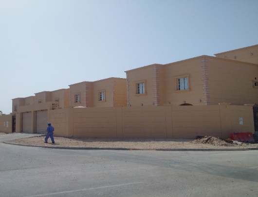 A row of beige, two-story houses under construction behind a beige wall, with a person walking on the sidewalk in front.
