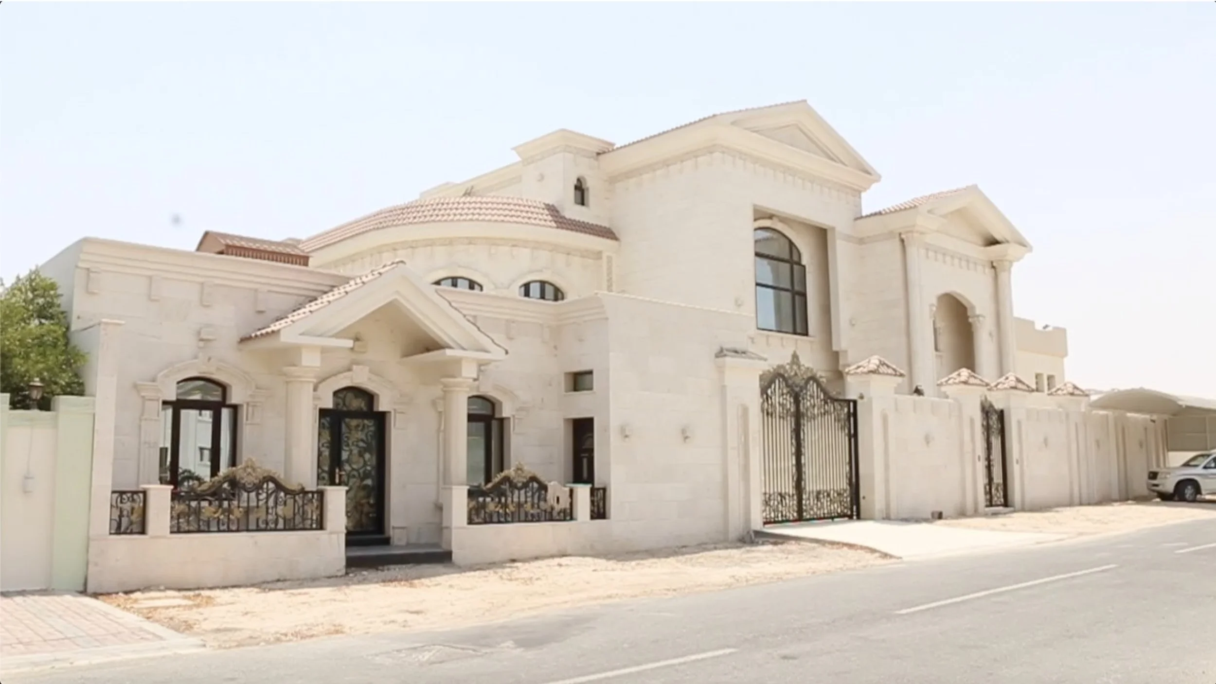 Large cream-colored mansion with multiple levels, arched windows, and an ornate wrought iron gate