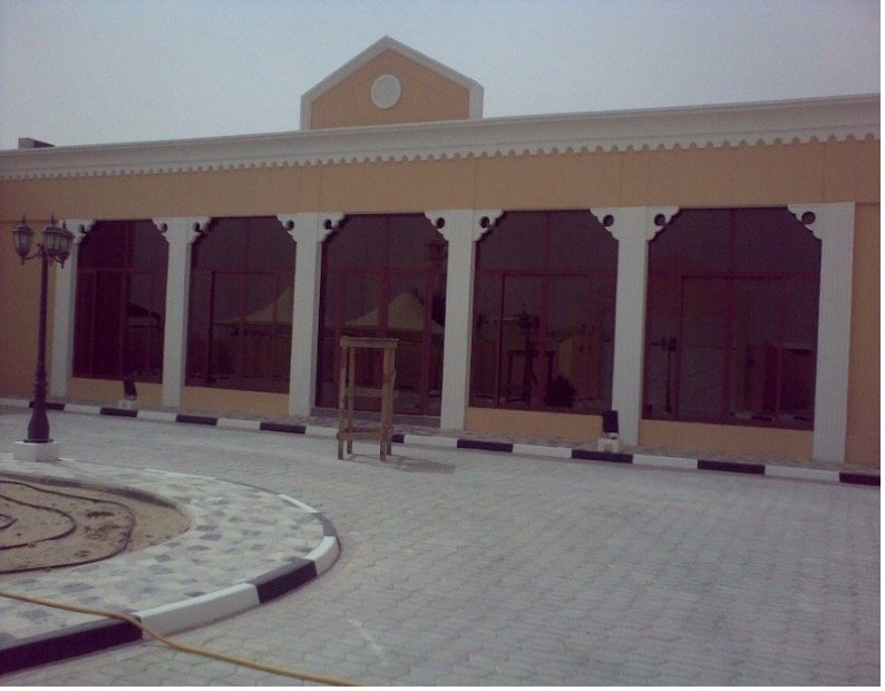 A building with large glass windows and tan walls, featuring a triangular roof with a circular window at the top. There is a paved courtyard in front with a lamp post, a small tree without leaves, and a curb painted with black and white stripes.