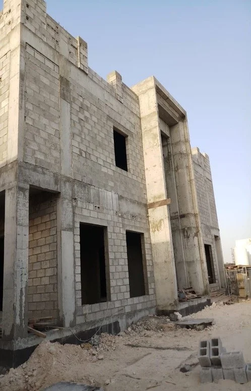 Concrete block building under construction with multiple empty window and door openings, construction debris on the ground, and a clear sky.