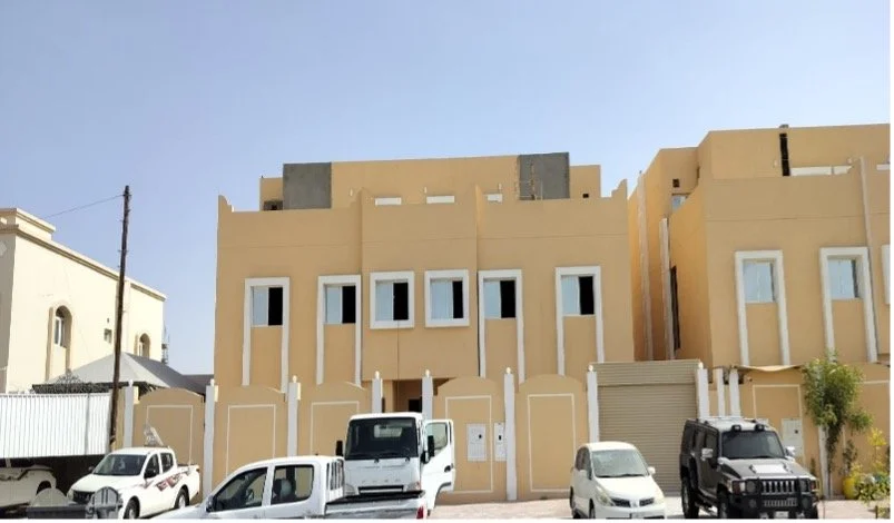 Street view of a beige-colored residential building with multiple windows, parked cars in front, including SUVs and a van, under a clear blue sky.