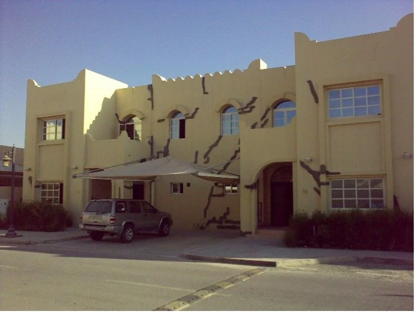 A yellow stucco building with dark brown cracks painted on the exterior walls, two stories, with multiple windows, a small balcony, and an entrance door. There is a parked SUV under a beige awning, and the building is situated on a paved street with a yellow parking curb.