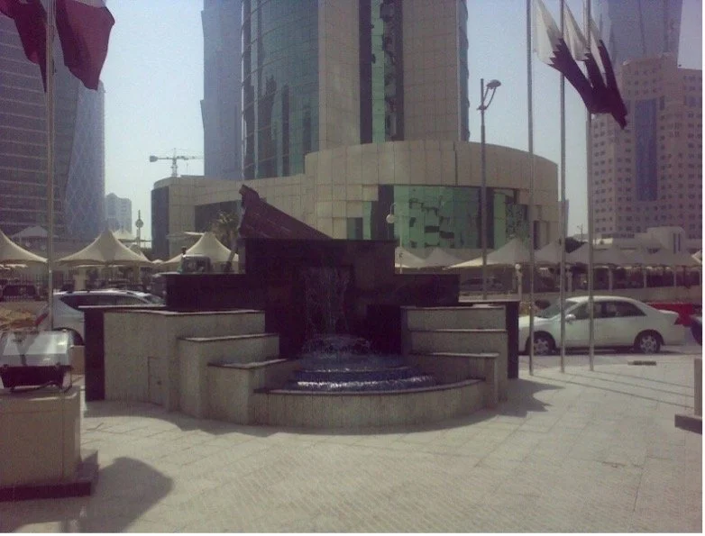 Urban plaza with modern building, water fountain, and cars parked nearby.