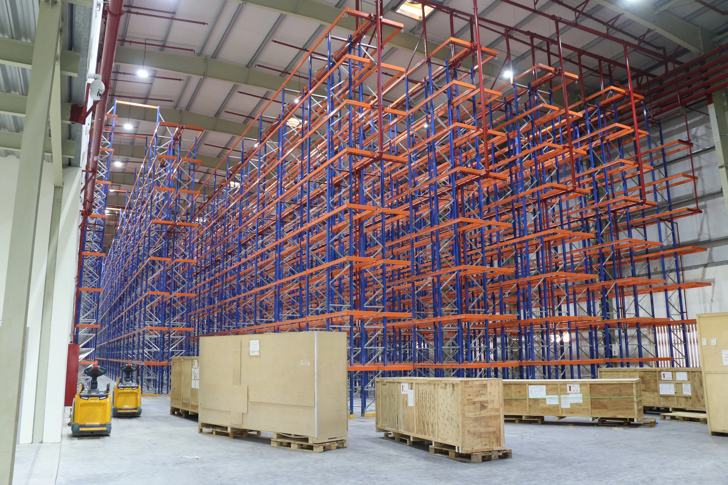 Large warehouse with blue and orange racking to store goods, some wooden crates, and two yellow pallet jacks on the floor.