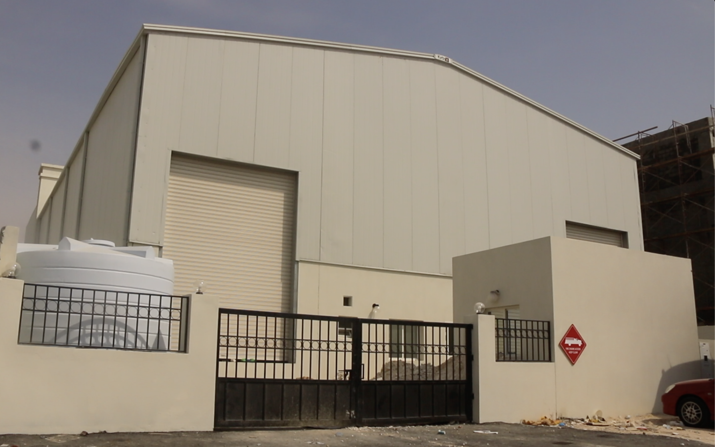 Large industrial building with a white exterior, metal rolling doors, a water tank, and a black gate in front with a warning sign. Clear sky background.