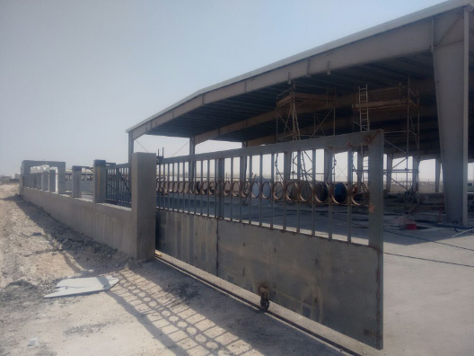 Construction site with scaffolding and a partially built structure behind a metal gate and fence, on a dusty ground under a clear sky.