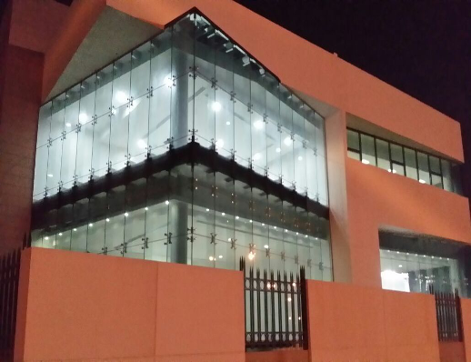 Modern building with large glass windows and illuminated interior at night, fenced by a metal gate.