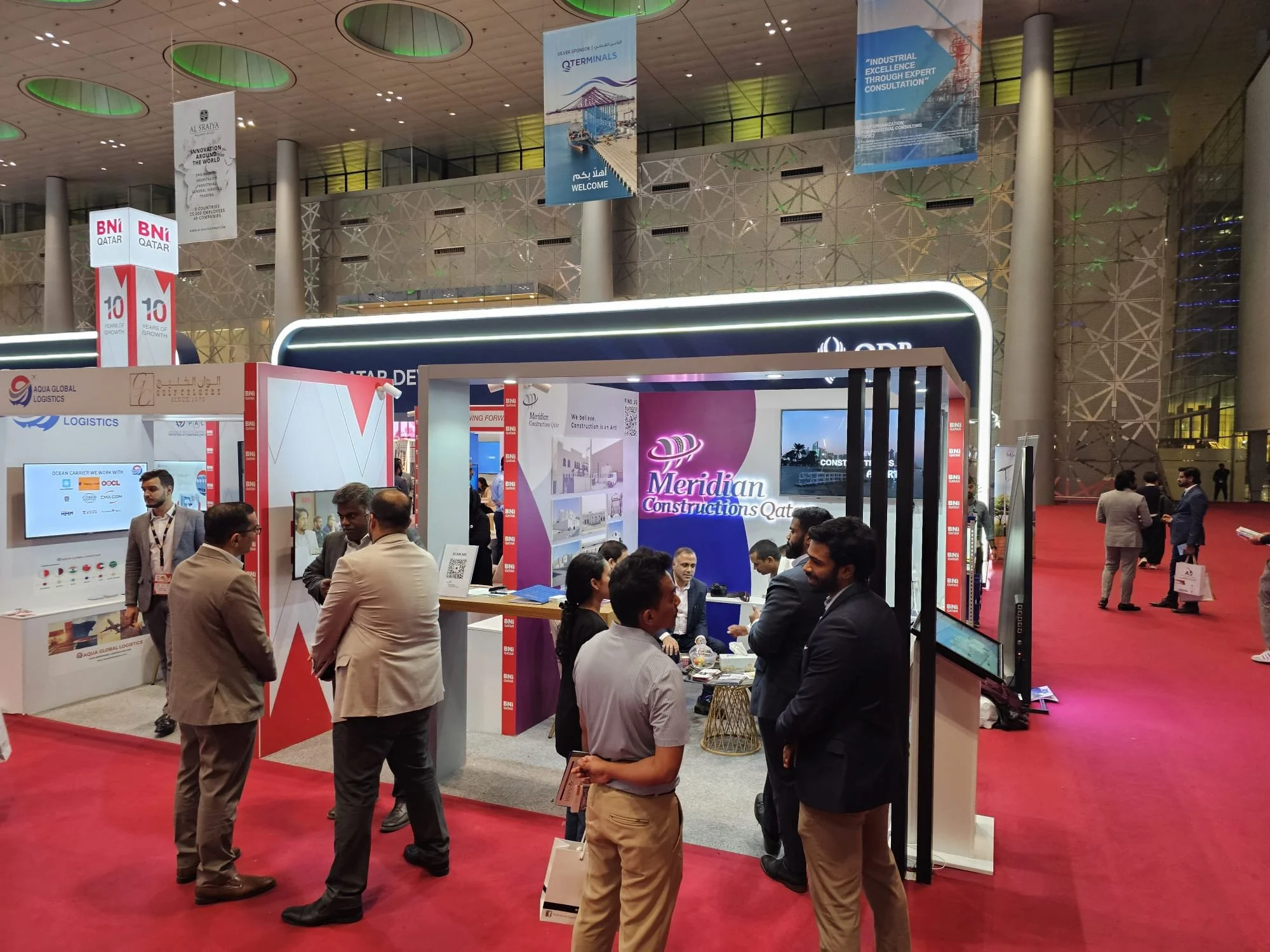 Trade show booth with people talking, signage displaying 'Meridian Constructions Qatar' and banners hanging from the ceiling in a large, modern hall.