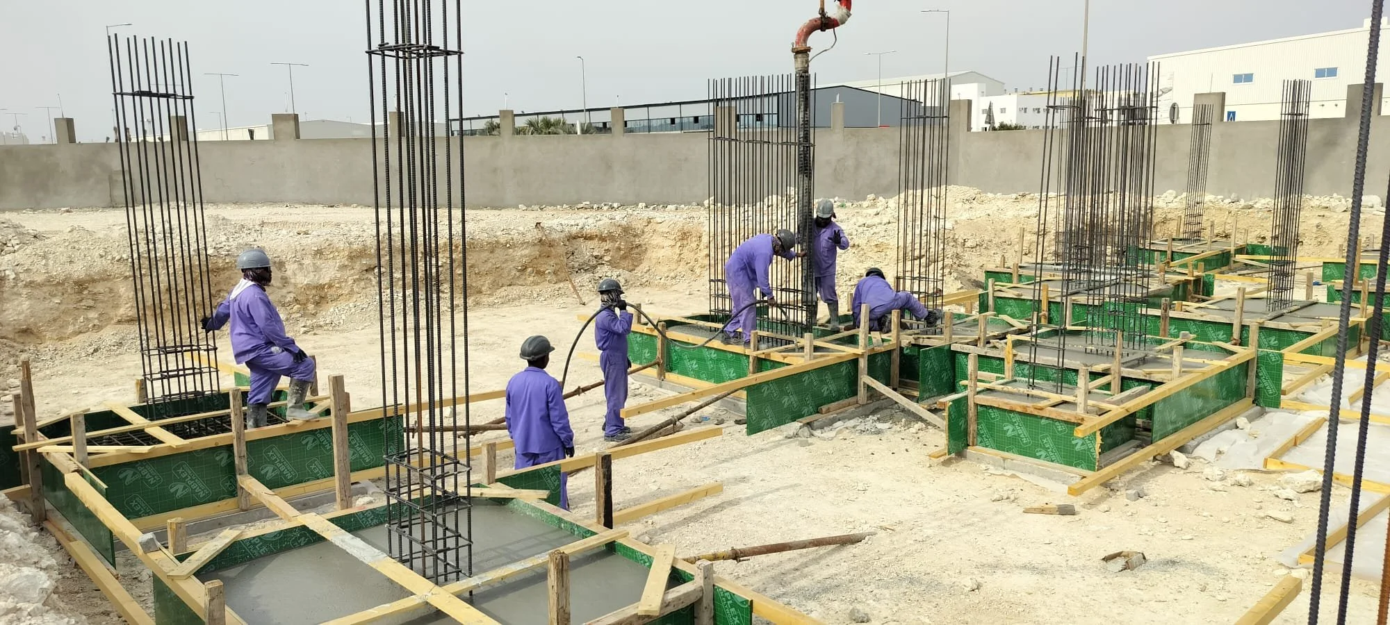Construction workers wearing purple uniforms and safety helmets working on building foundations with rebar and wooden formwork at a construction site.