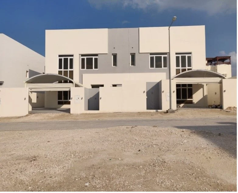 Modern white residential building with two symmetrical entrances and a partially covered patio, situated on a dirt lot under a blue sky.
