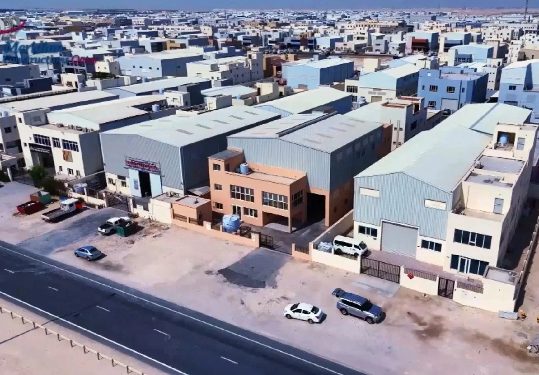 Aerial view of a densely packed industrial or commercial area with warehouses, factories, and parking lot with several cars. The buildings have metal roofs and are arranged in a grid pattern.