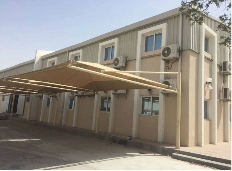 The exterior of a beige building with multiple windows, air conditioning units, and a large retractable beige awning extended over a parking area.